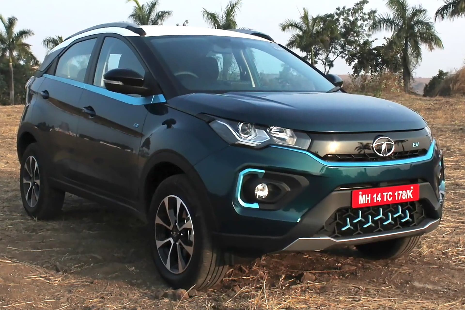 Teal Tata Nexon EV electric SUV with a white roof, front three-quarter view, parked on dry grass