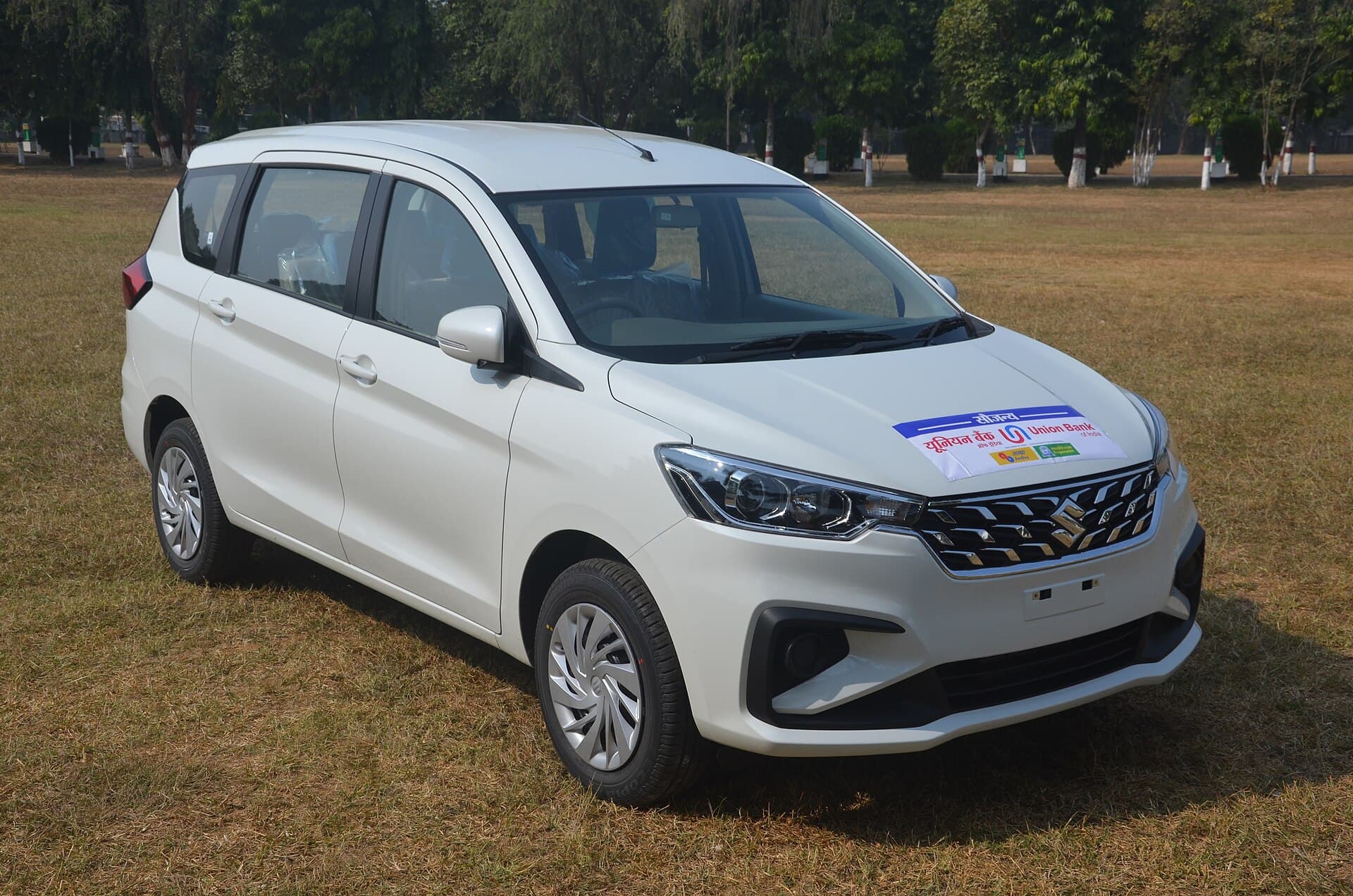 White Maruti Suzuki Ertiga seven-seater MUV, front three-quarter view, parked on a grass field